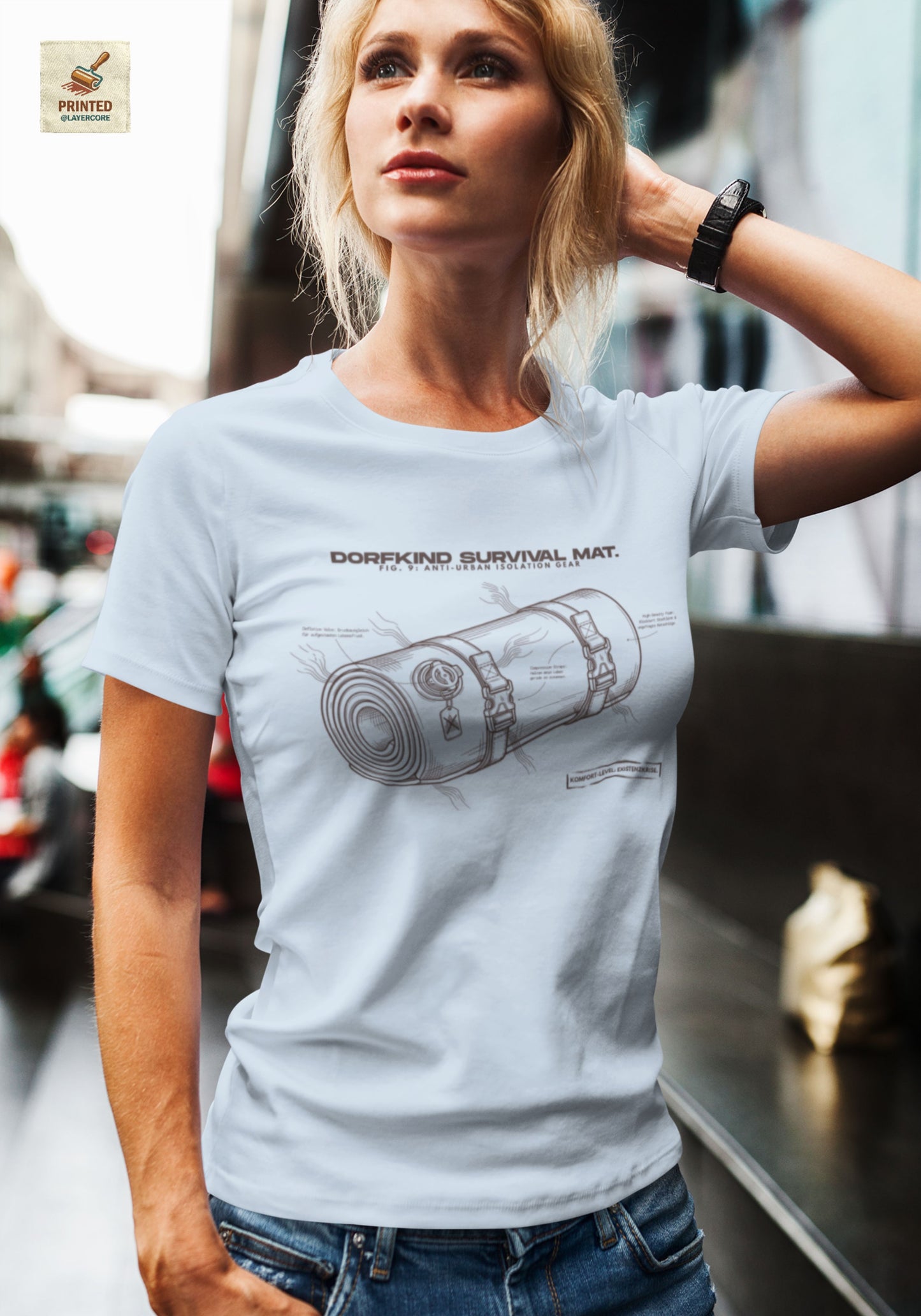 Technische Blueprint-Zeichnung einer Isomatte mit ironischen Texten zum Dorfleben auf einem hellblauen T-Shirt, getragen von einer jungen Frau. Anti-Gorpcore Design.