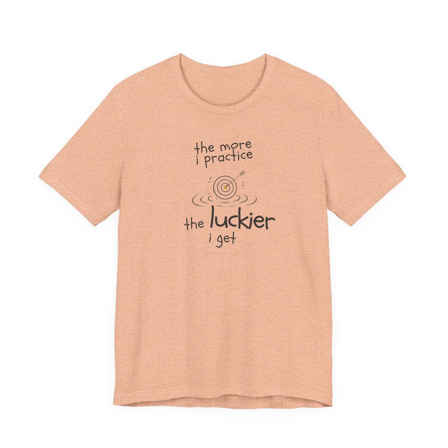 Minimalist motivational T-shirt design shown on a peach coloured shirt: hand-drawn cream lettering reads “the more i practice” above a sketched bullseye with an arrow and small mustard center, ripple lines underneath, then larger text reads “the luckier i get” with tiny mono text below that says “repetition > luck.”