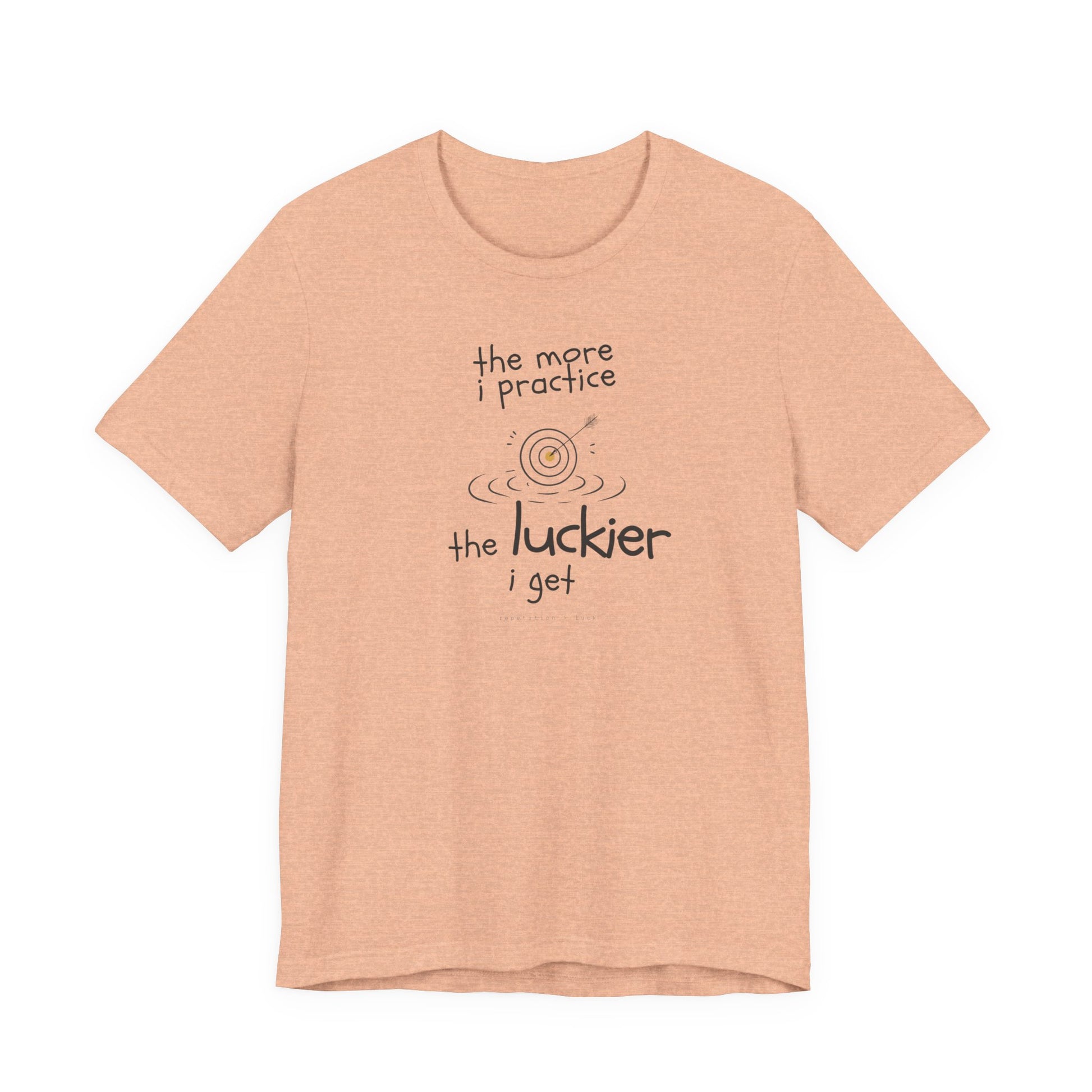 Minimalist motivational T-shirt design shown on a peach coloured shirt: hand-drawn cream lettering reads “the more i practice” above a sketched bullseye with an arrow and small mustard center, ripple lines underneath, then larger text reads “the luckier i get” with tiny mono text below that says “repetition > luck.”