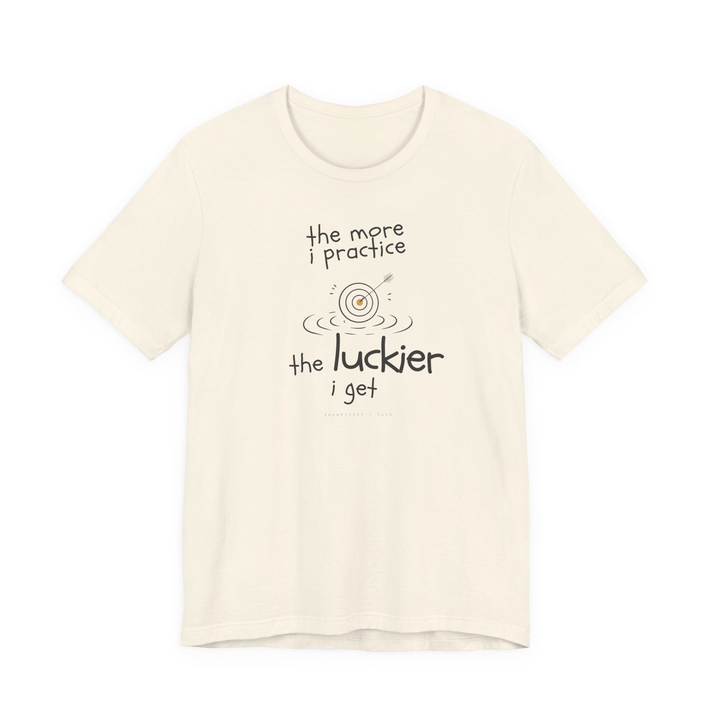 Minimalist motivational T-shirt design shown on a natural coloured shirt: hand-drawn cream lettering reads “the more i practice” above a sketched bullseye with an arrow and small mustard center, ripple lines underneath, then larger text reads “the luckier i get” with tiny mono text below that says “repetition > luck.”
