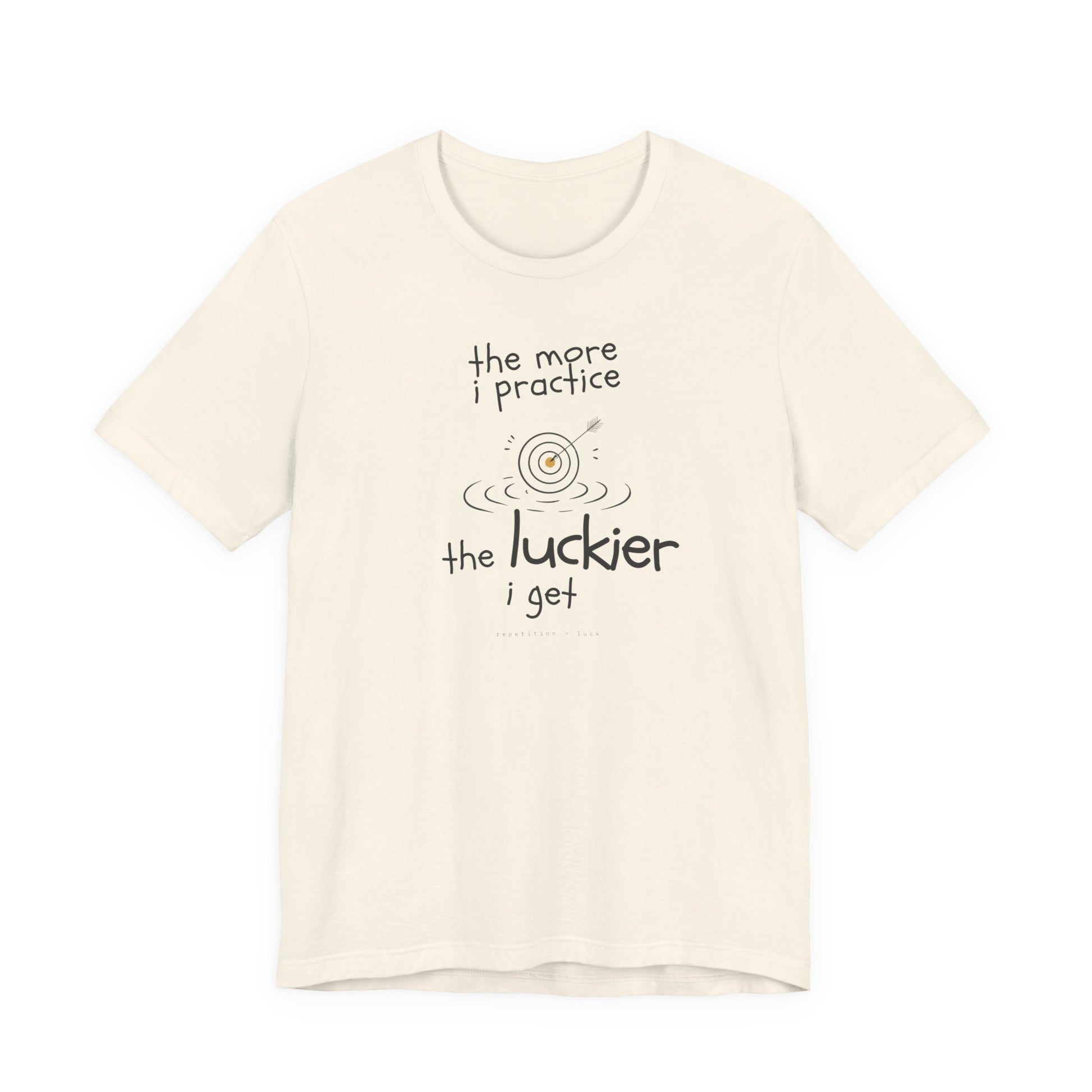 Minimalist motivational T-shirt design shown on a natural coloured shirt: hand-drawn cream lettering reads “the more i practice” above a sketched bullseye with an arrow and small mustard center, ripple lines underneath, then larger text reads “the luckier i get” with tiny mono text below that says “repetition > luck.”