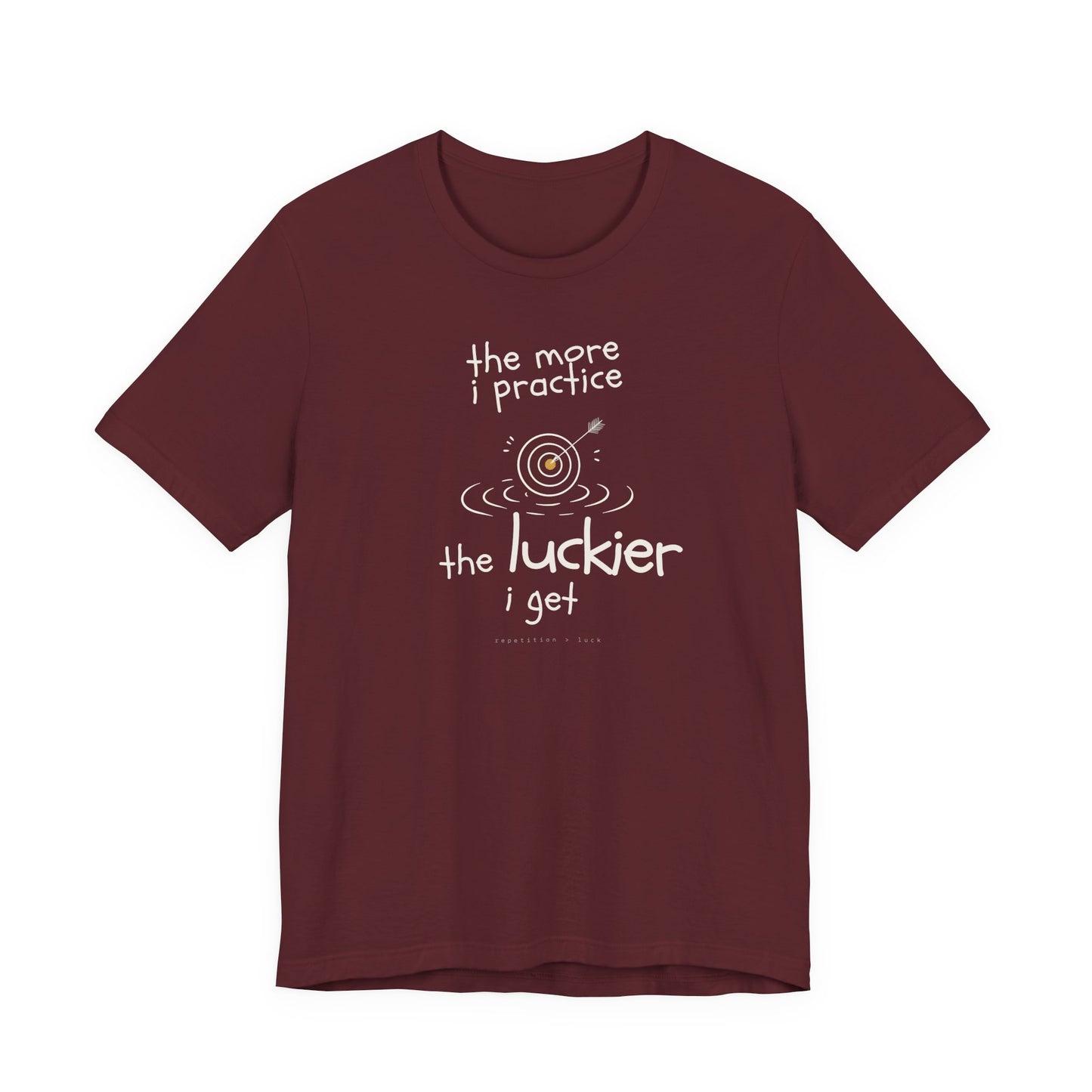 Minimalist motivational T-shirt design shown on a maroon coloured shirt: hand-drawn cream lettering reads “the more i practice” above a sketched bullseye with an arrow and small mustard center, ripple lines underneath, then larger text reads “the luckier i get” with tiny mono text below that says “repetition > luck.”