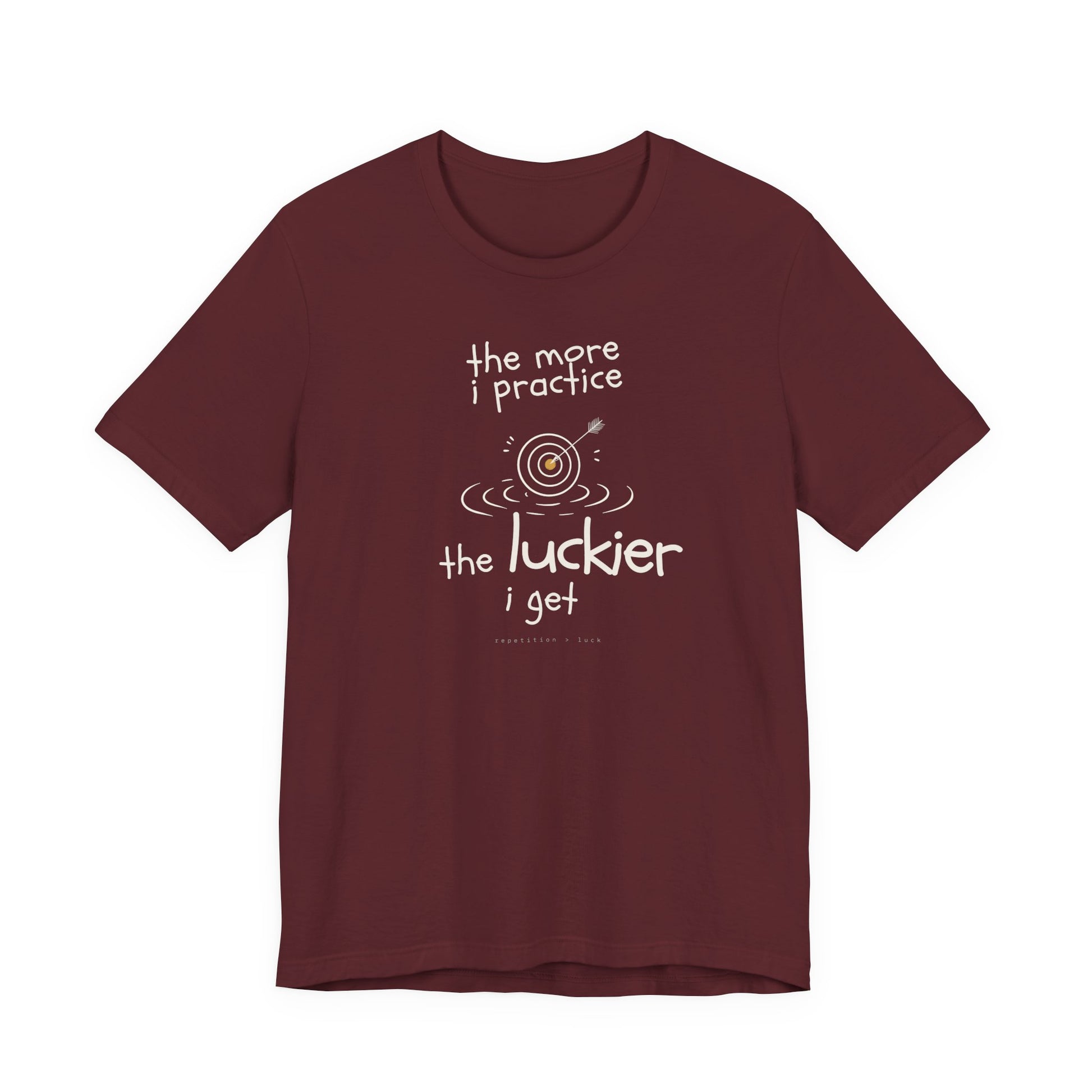 Minimalist motivational T-shirt design shown on a maroon coloured shirt: hand-drawn cream lettering reads “the more i practice” above a sketched bullseye with an arrow and small mustard center, ripple lines underneath, then larger text reads “the luckier i get” with tiny mono text below that says “repetition > luck.”