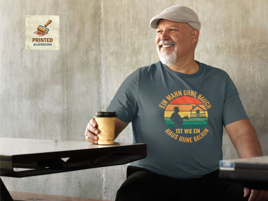 Ein Mann mittleren Alters mit einem sichtbaren Bauch trägt das blaue "Mann ohne Bauch" T-Shirt in einer entspannten Street-Atmosphäre. Das Retro-Sonnenuntergangs-Design in warmen Orange- und Gelbtönen kommt auf dem Stoff gut zur Geltung. Das Shirt aus Bella Canvas 3001 Baumwolle sitzt locker. Der Spruch zelebriert auf humorvolle, selbstironische Weise und passt perfekt zu geselligen Runden wie Stammtischen oder Grillabenden im deutschsprachigen Raum. Es ist ein Statement für Gelassenheit und Genuss.