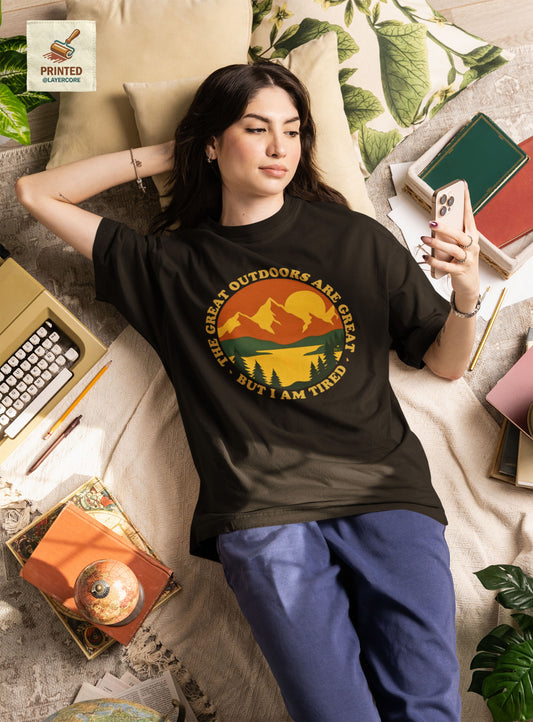 Retro 70s Berglandschaft schwarzes T-Shirt, getargen von einer jungen Frau, welche auf einem bett umringt von Gegenständen liegt, mit "The Great Outdoors are great but I am tired" Schriftzug.