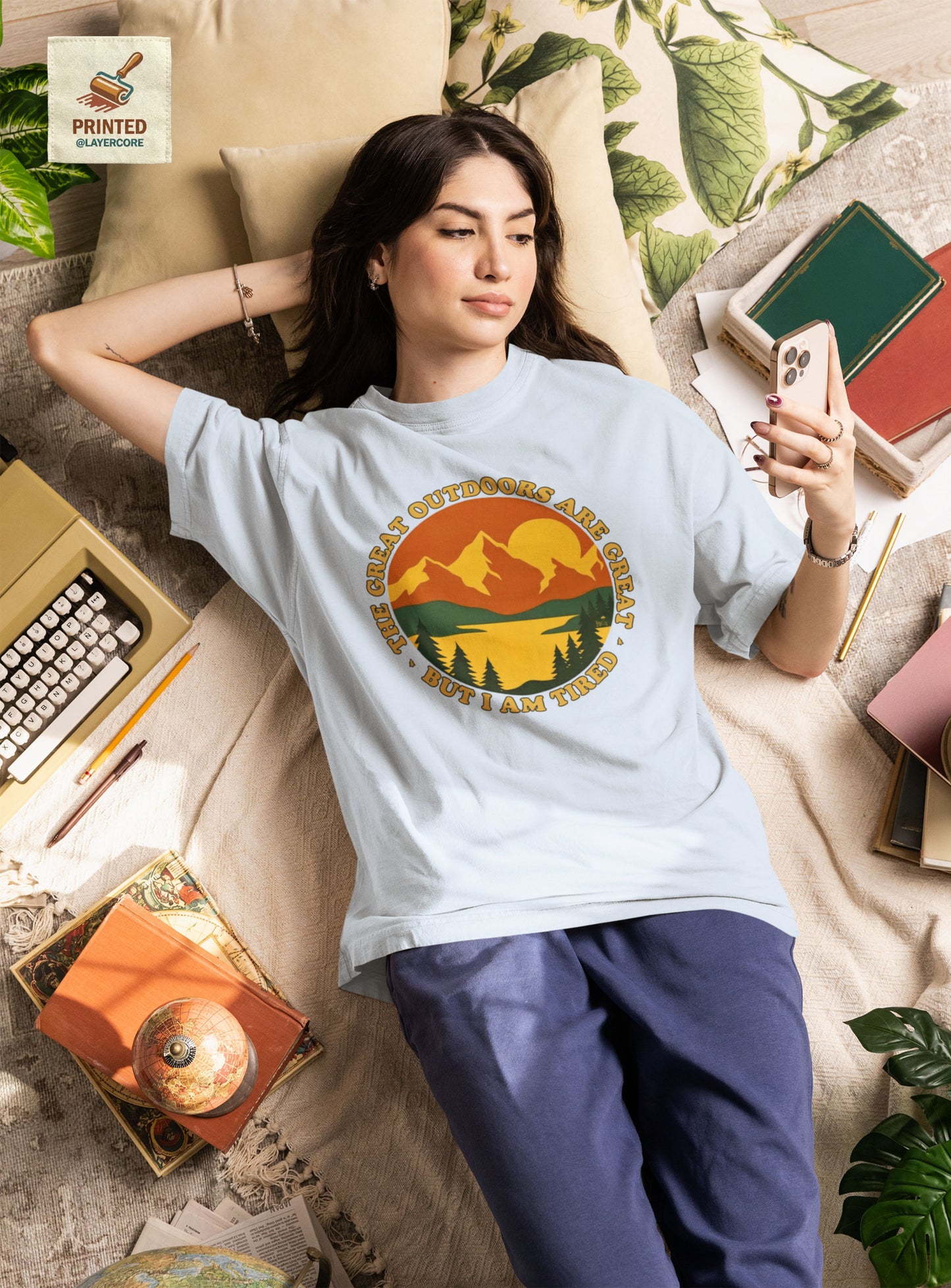 Retro 70s Berglandschaft hellblaues T-Shirt, getargen von einer jungen Frau, welche auf einem bett umringt von Gegenständen liegt, mit "The Great Outdoors are great but I am tired" Schriftzug.
