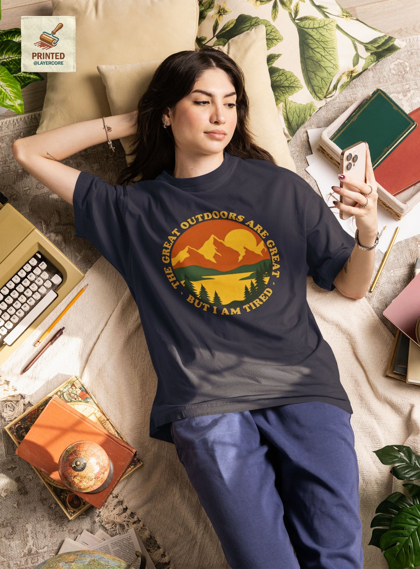 Retro 70s Berglandschaft navy blaues T-Shirt, getargen von einer jungen Frau, welche auf einem bett umringt von Gegenständen liegt, mit "The Great Outdoors are great but I am tired" Schriftzug.