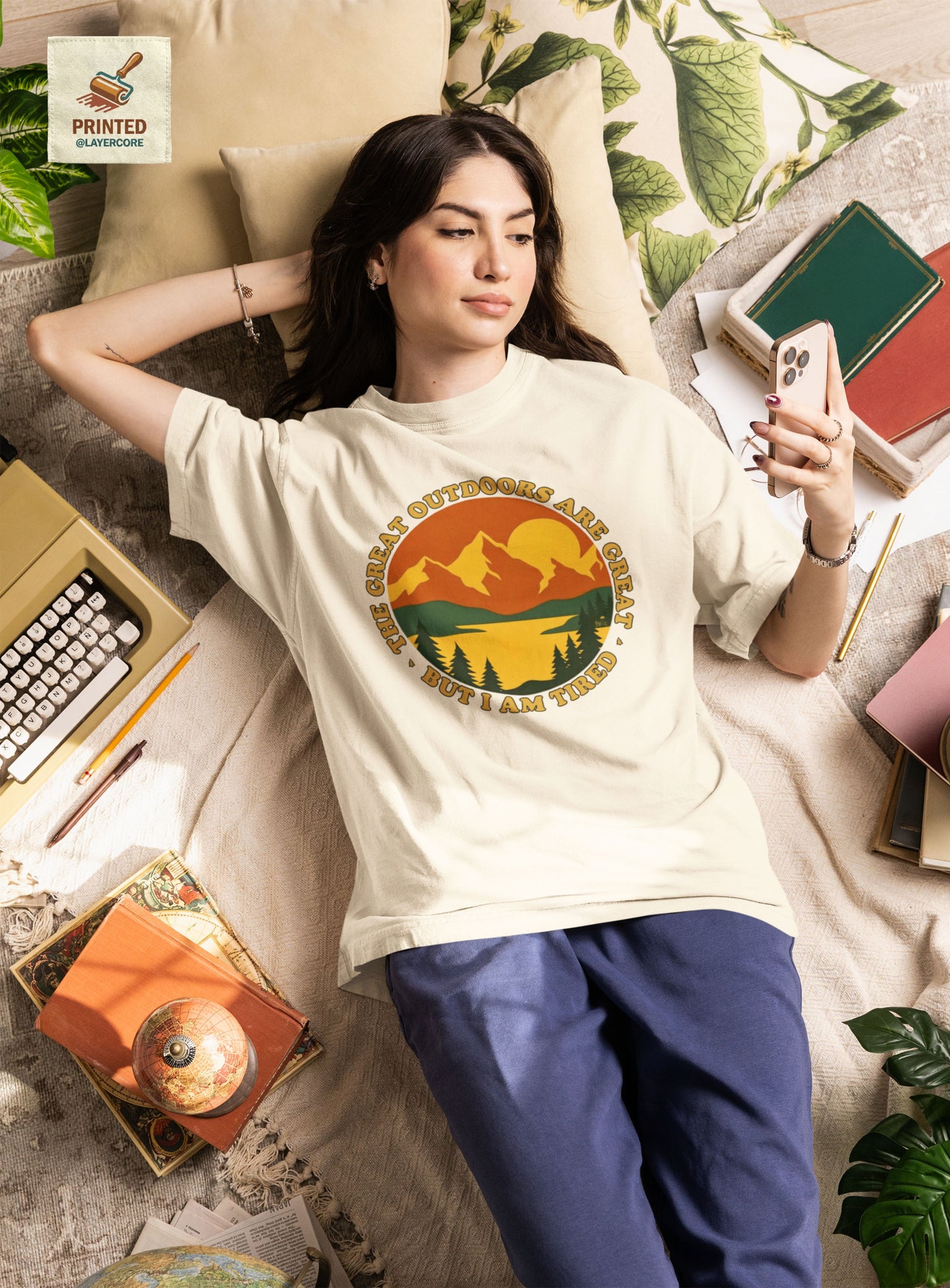 Retro 70s Berglandschaft natural farbenes T-Shirt, getargen von einer jungen Frau, welche auf einem bett umringt von Gegenständen liegt, mit "The Great Outdoors are great but I am tired" Schriftzug.
