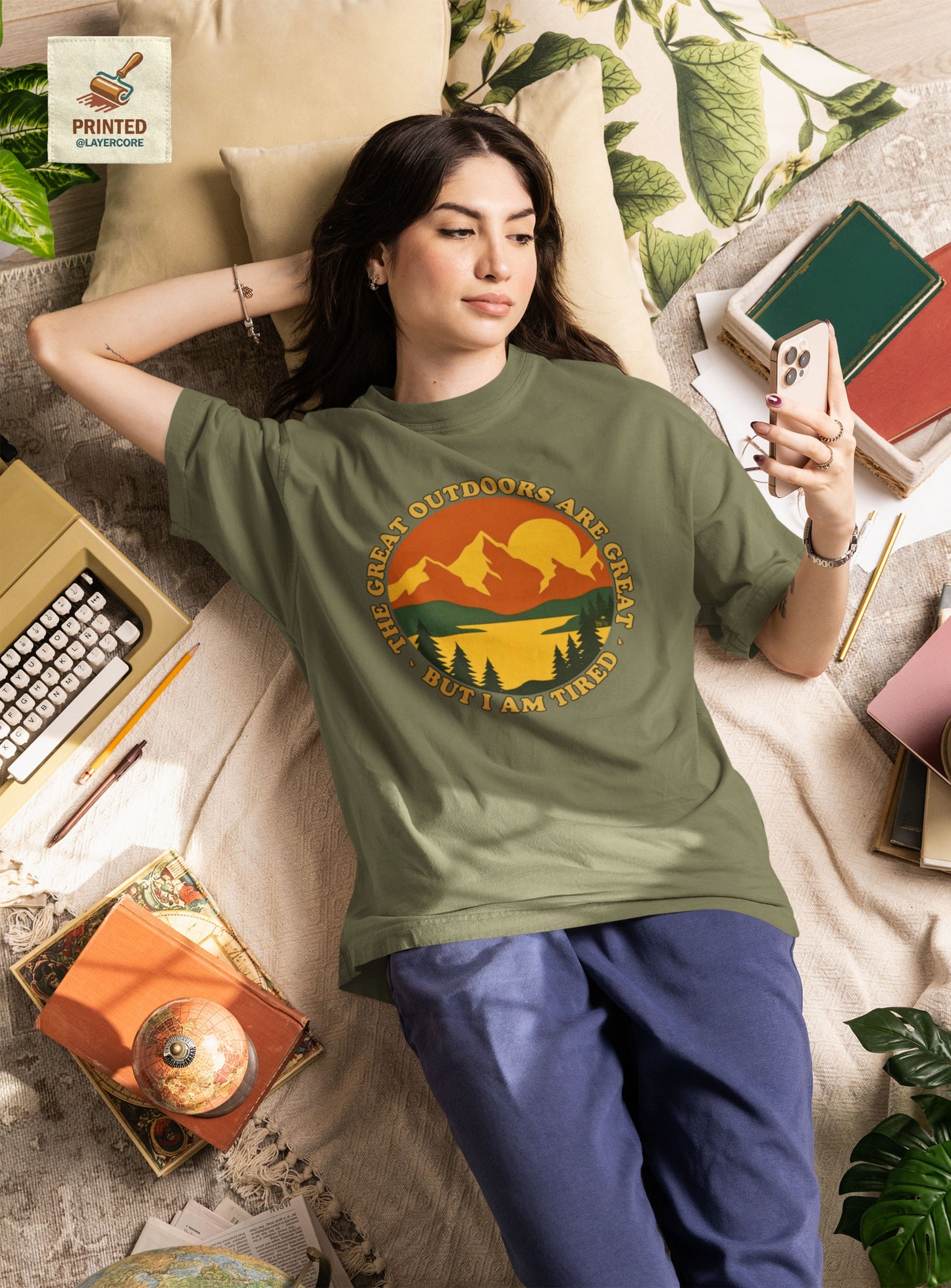 Retro 70s Berglandschaft military grünes T-Shirt, getargen von einer jungen Frau, welche auf einem bett umringt von Gegenständen liegt, mit "The Great Outdoors are great but I am tired" Schriftzug.