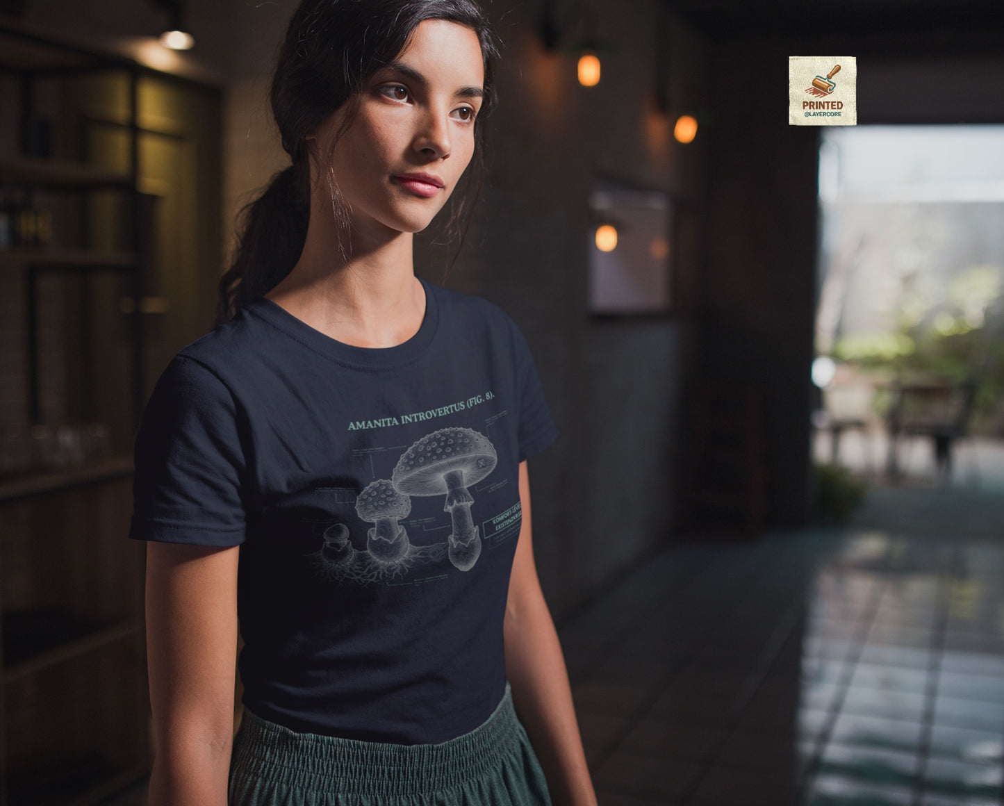 Vintage-inspiriertes blaues T-Shirt mit einer detaillierten Kupferstich-Illustration von Fliegenpilzen (Amanita muscaria), getragen von einer jungen Frau. Das Design trägt den ironischen lateinischen Titel "AMANITA INTROVERTUS" und beschreibt anatomische Merkmale mit humorvollen denglischen Texten über soziale Erschöpfung, wie "Komfort-Level: Existenzkrise" und "Hidden Anxiety". Ein hochwertiges Dark Academia Graphic Tee für Introvertierte und Studenten.