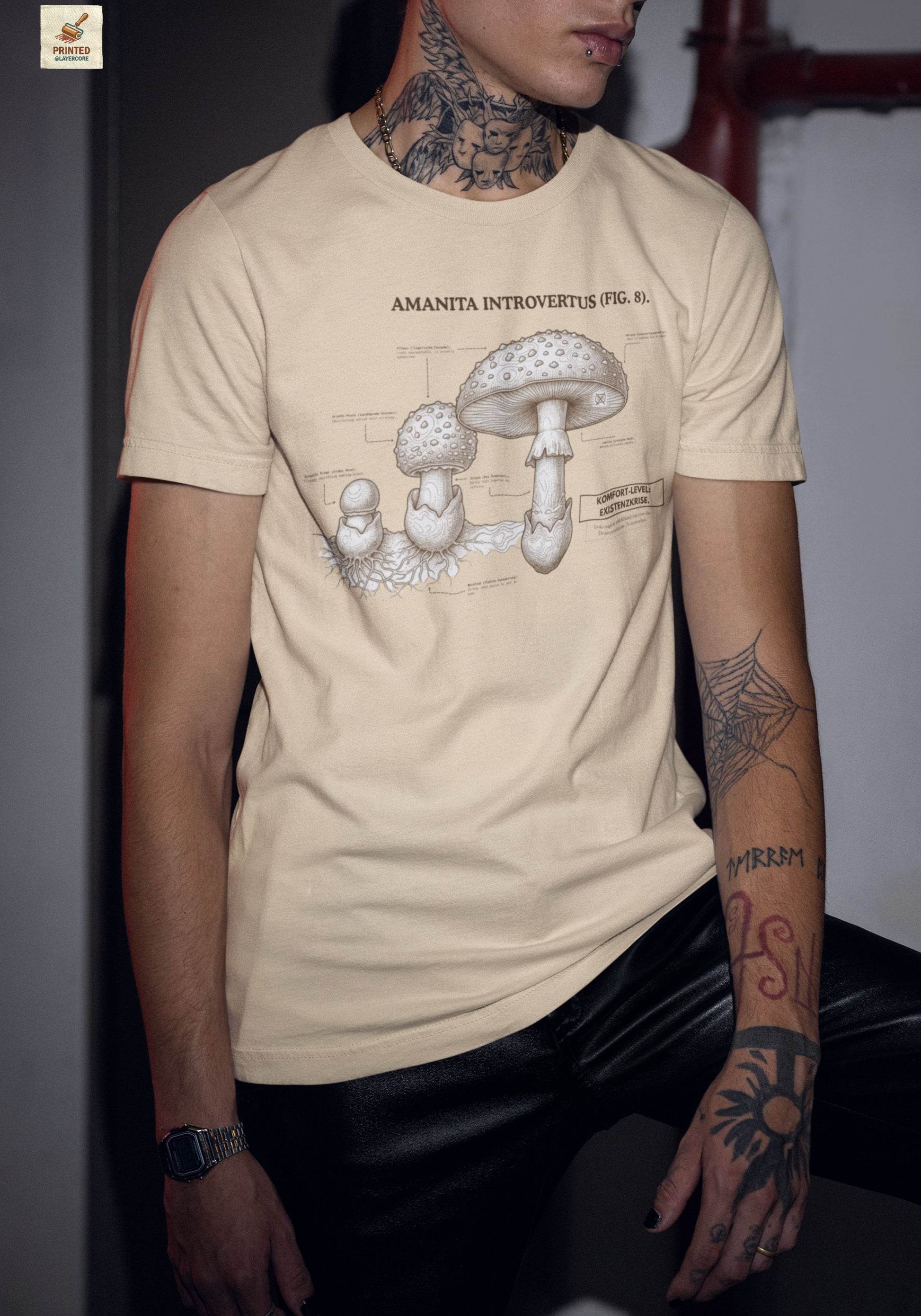Vintage-inspiriertes natur farbenes T-Shirt mit einer detaillierten Kupferstich-Illustration von Fliegenpilzen (Amanita muscaria), getragen von einem jungen Mann. Das Design trägt den ironischen lateinischen Titel "AMANITA INTROVERTUS" und beschreibt anatomische Merkmale mit humorvollen denglischen Texten über soziale Erschöpfung, wie "Komfort-Level: Existenzkrise" und "Hidden Anxiety". Ein hochwertiges Dark Academia Graphic Tee für Introvertierte und Studenten.