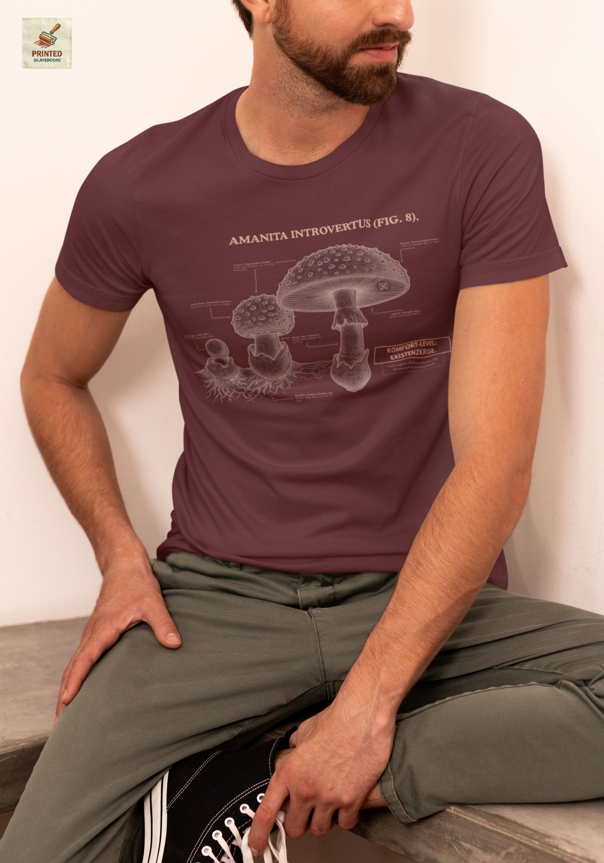 Vintage-inspiriertes rotes T-Shirt mit einer detaillierten Kupferstich-Illustration von Fliegenpilzen (Amanita muscaria), getragen von einem jungen Mann. Das Design trägt den ironischen lateinischen Titel "AMANITA INTROVERTUS" und beschreibt anatomische Merkmale mit humorvollen denglischen Texten über soziale Erschöpfung, wie "Komfort-Level: Existenzkrise" und "Hidden Anxiety". Ein hochwertiges Dark Academia Graphic Tee für Introvertierte und Studenten.