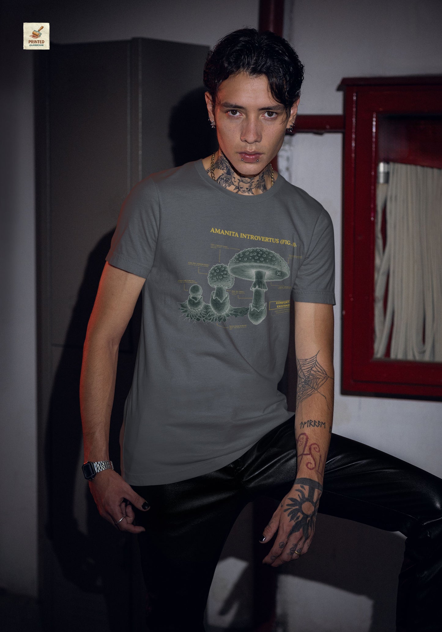 Vintage-inspiriertes asphalt farbenes T-Shirt mit einer detaillierten Kupferstich-Illustration von Fliegenpilzen (Amanita muscaria), getragen von einem jungen Mann. Das Design trägt den ironischen lateinischen Titel "AMANITA INTROVERTUS" und beschreibt anatomische Merkmale mit humorvollen denglischen Texten über soziale Erschöpfung, wie "Komfort-Level: Existenzkrise" und "Hidden Anxiety". Ein hochwertiges Dark Academia Graphic Tee für Introvertierte und Studenten.