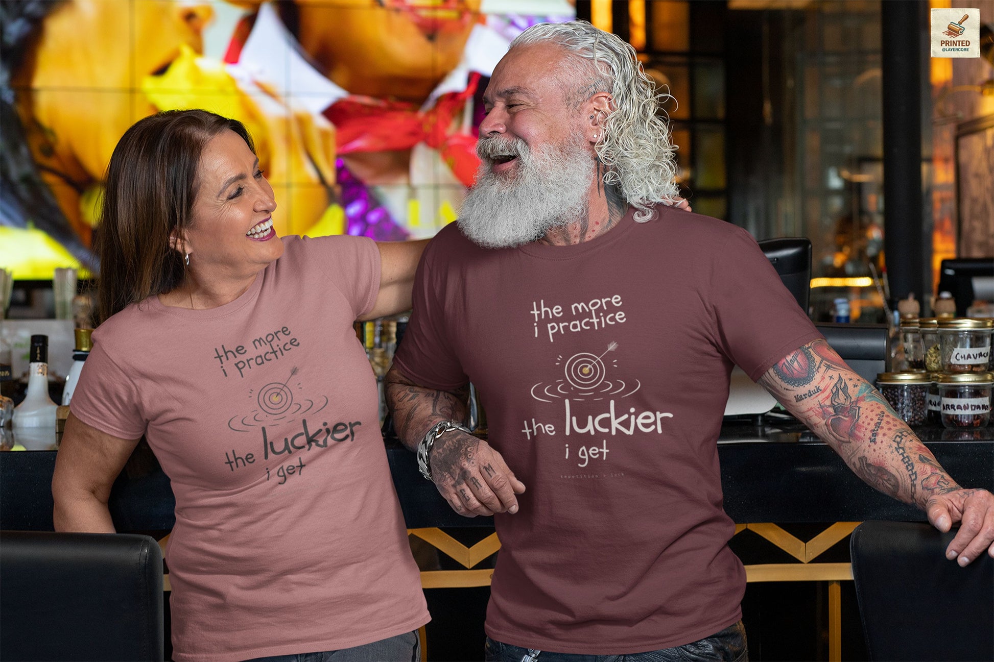 Minimalist motivational T-shirt design shown on a maroon and a peach coloured shirt: hand-drawn cream lettering reads “the more i practice” above a sketched bullseye with an arrow and small mustard center, ripple lines underneath, then larger text reads “the luckier i get” with tiny mono text below that says “repetition > luck.”