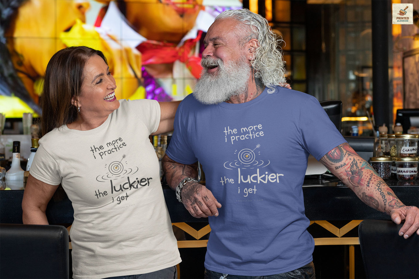 Minimalist motivational T-shirt design shown on a light blue and a natural coloured shirt: hand-drawn cream lettering reads “the more i practice” above a sketched bullseye with an arrow and small mustard center, ripple lines underneath, then larger text reads “the luckier i get” with tiny mono text below that says “repetition > luck.”