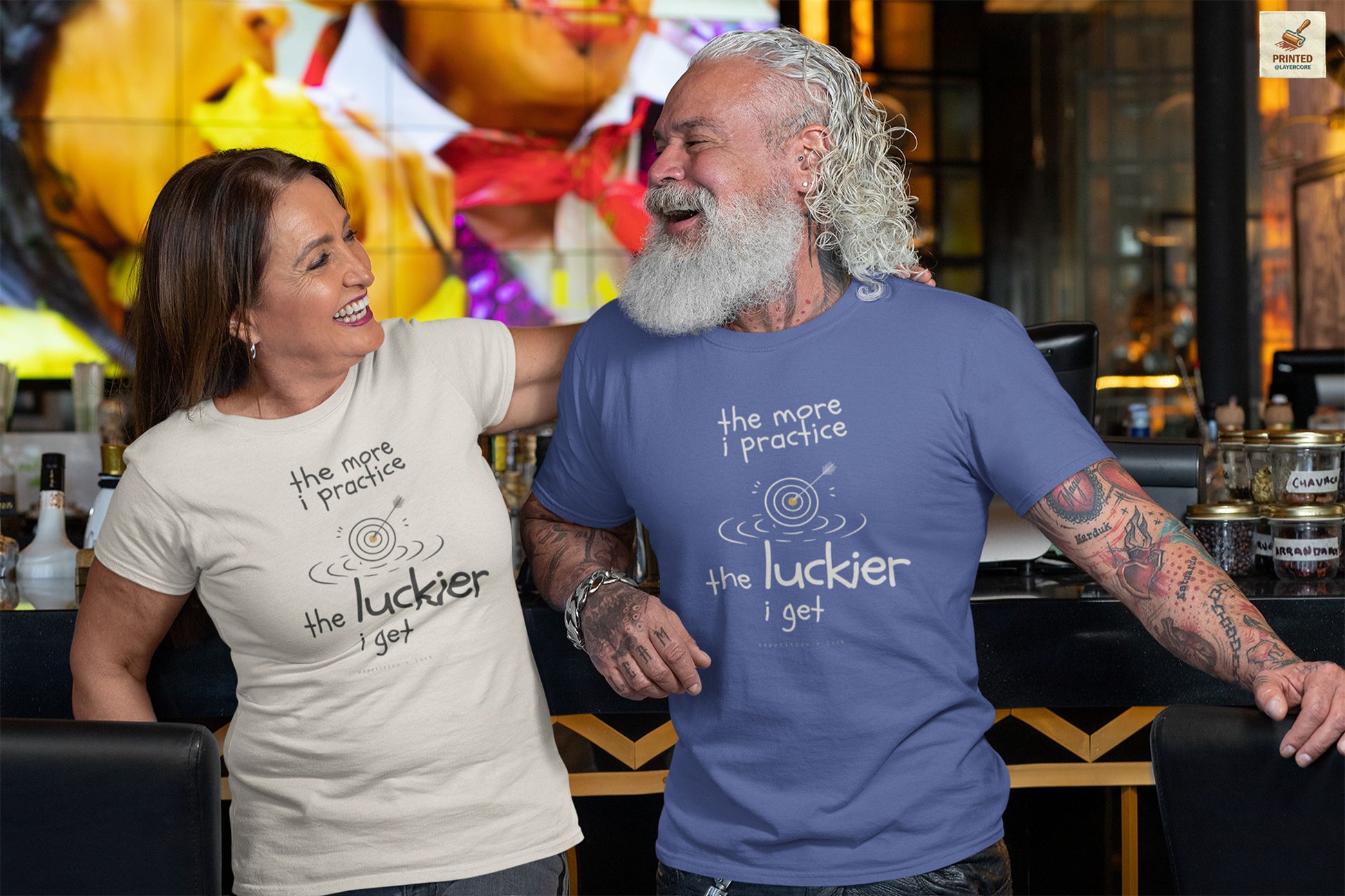 Minimalist motivational T-shirt design shown on a light blue and a natural coloured shirt: hand-drawn cream lettering reads “the more i practice” above a sketched bullseye with an arrow and small mustard center, ripple lines underneath, then larger text reads “the luckier i get” with tiny mono text below that says “repetition > luck.”