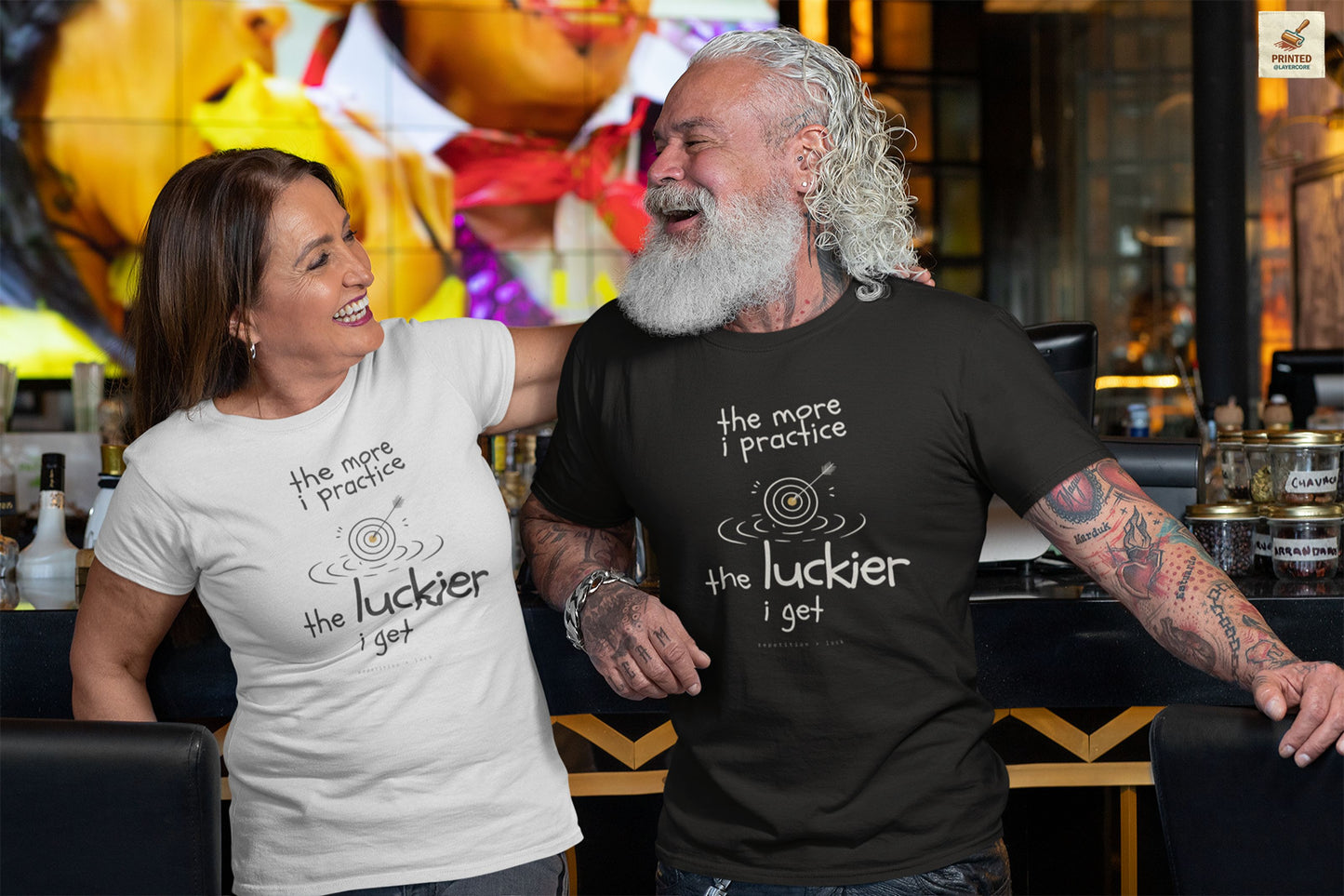 Minimalist motivational T-shirt design shown on a black and a white shirt: hand-drawn cream lettering reads “the more i practice” above a sketched bullseye with an arrow and small mustard center, ripple lines underneath, then larger text reads “the luckier i get” with tiny mono text below that says “repetition > luck.”