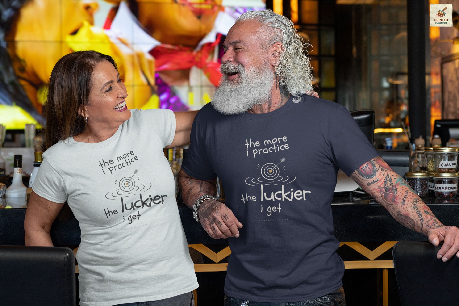 Minimalist motivational T-shirt design shown on a dark blue and a white shirt: hand-drawn cream lettering reads “the more i practice” above a sketched bullseye with an arrow and small mustard center, ripple lines underneath, then larger text reads “the luckier i get” with tiny mono text below that says “repetition > luck.”