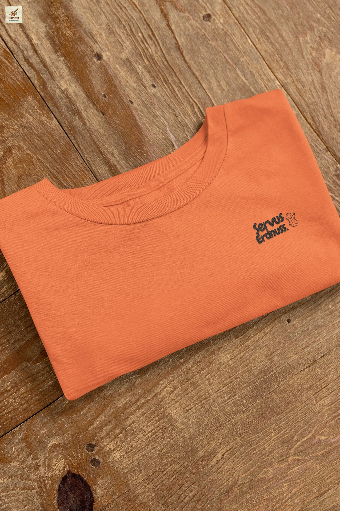 Servus Erdnuss Brustmotiv mit Dialekt-Text und kleiner Erdnussfigur auf orangenem Shirt