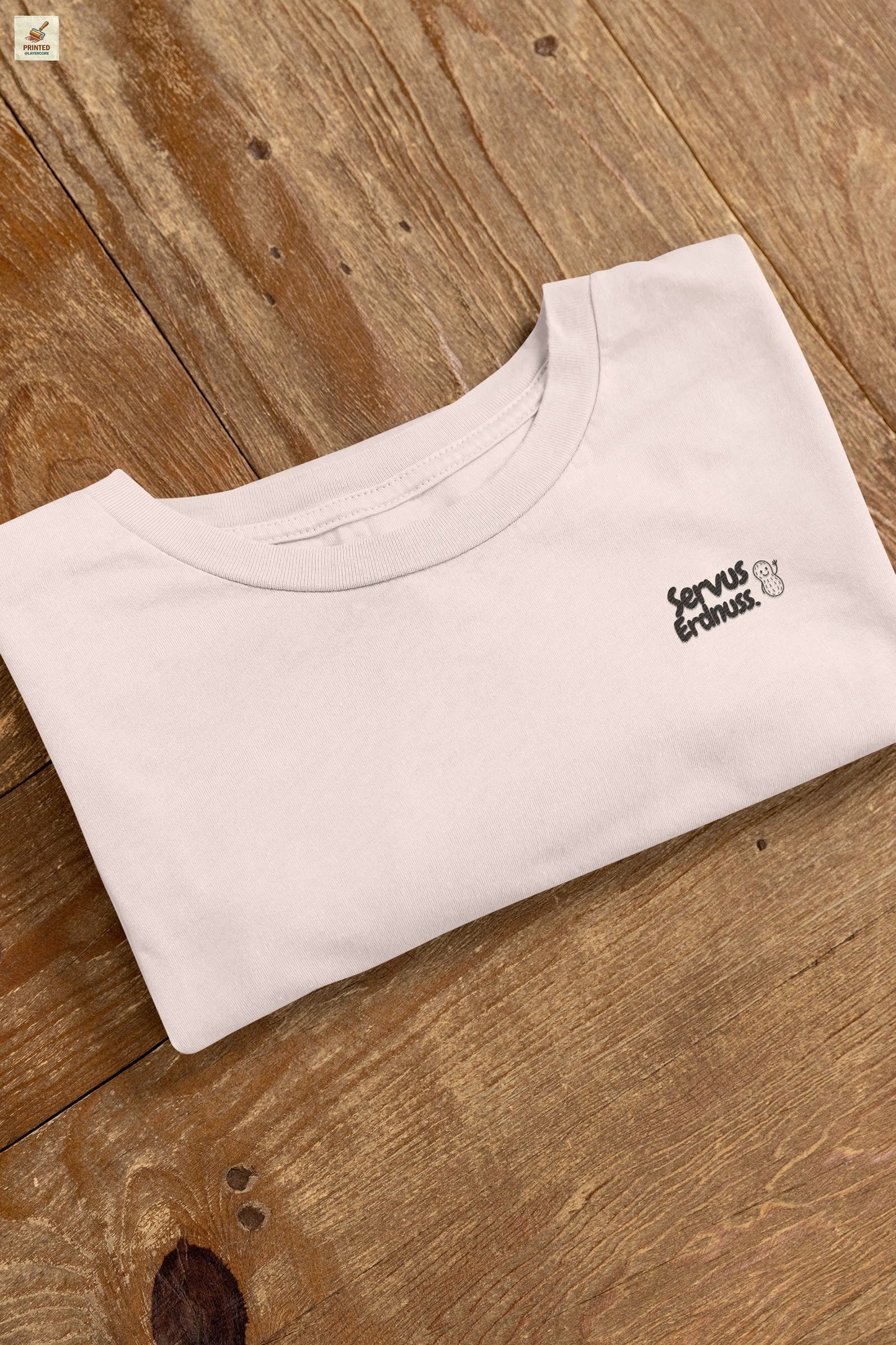 Servus Erdnuss Brustmotiv mit Dialekt-Text und kleiner Erdnussfigur auf soft pinkem Shirt