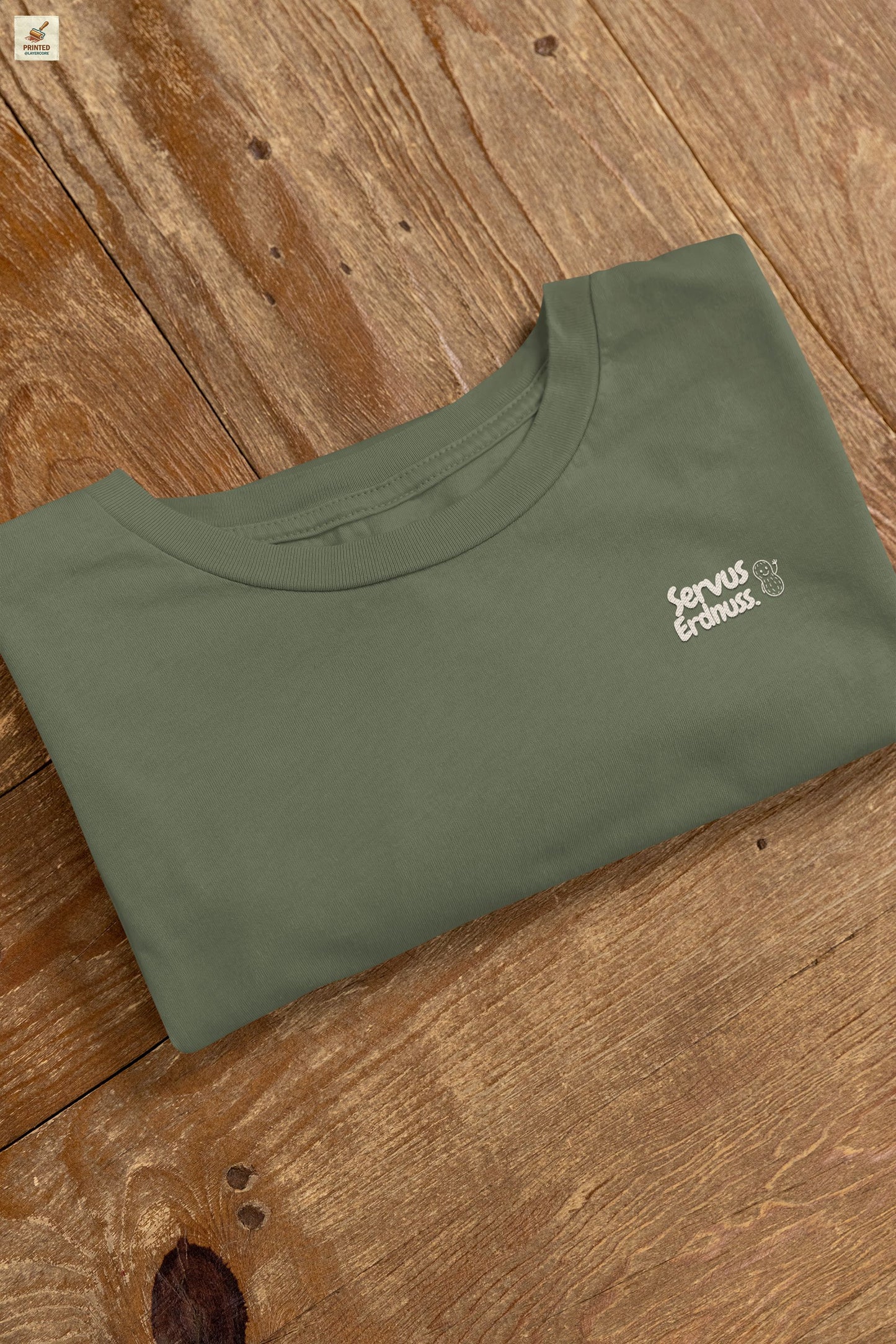 Servus Erdnuss Brustmotiv mit Dialekt-Text und kleiner Erdnussfigur auf grünem Shirt