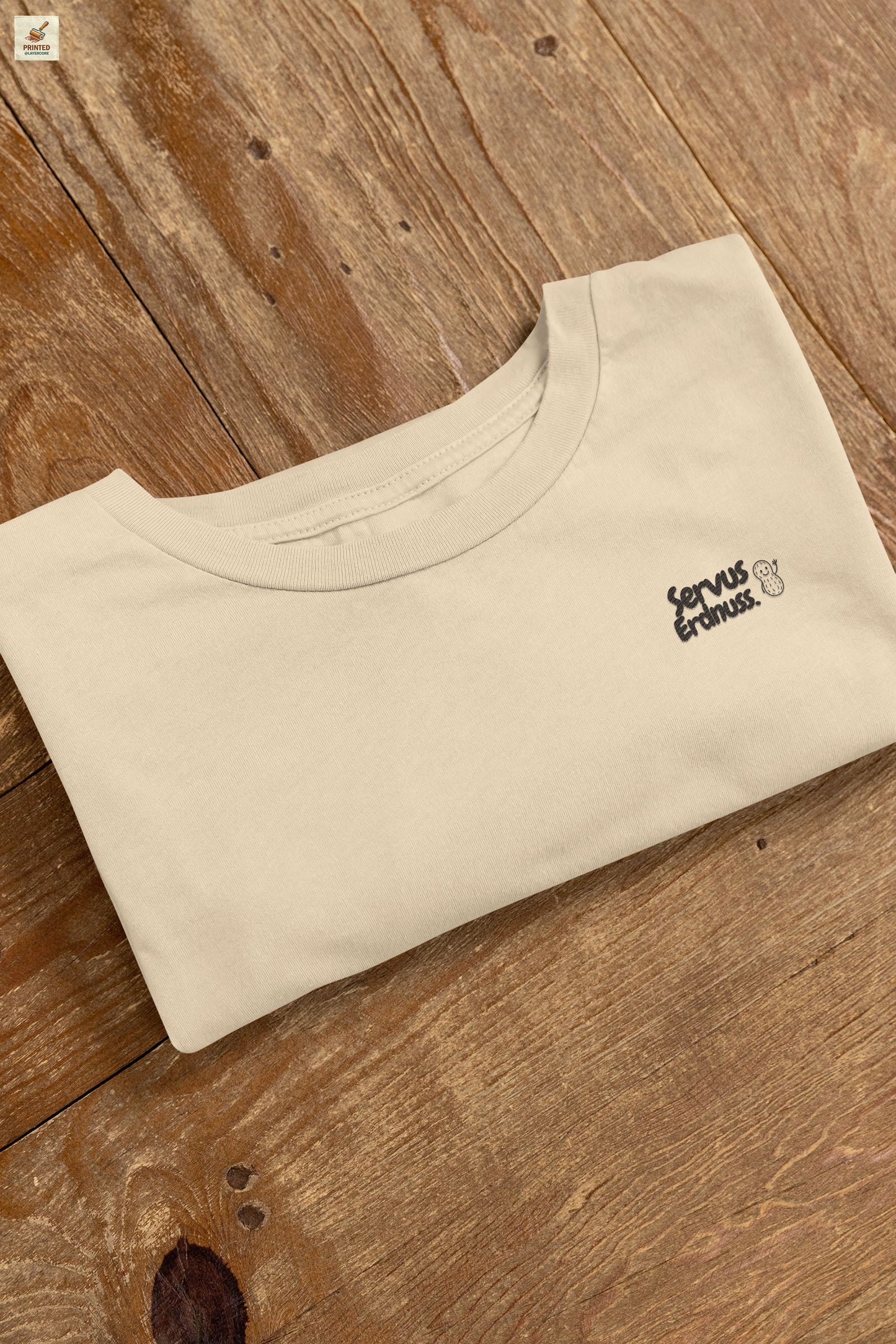 Servus Erdnuss Brustmotiv mit Dialekt-Text und kleiner Erdnussfigur auf naturell farbenem Shirt
