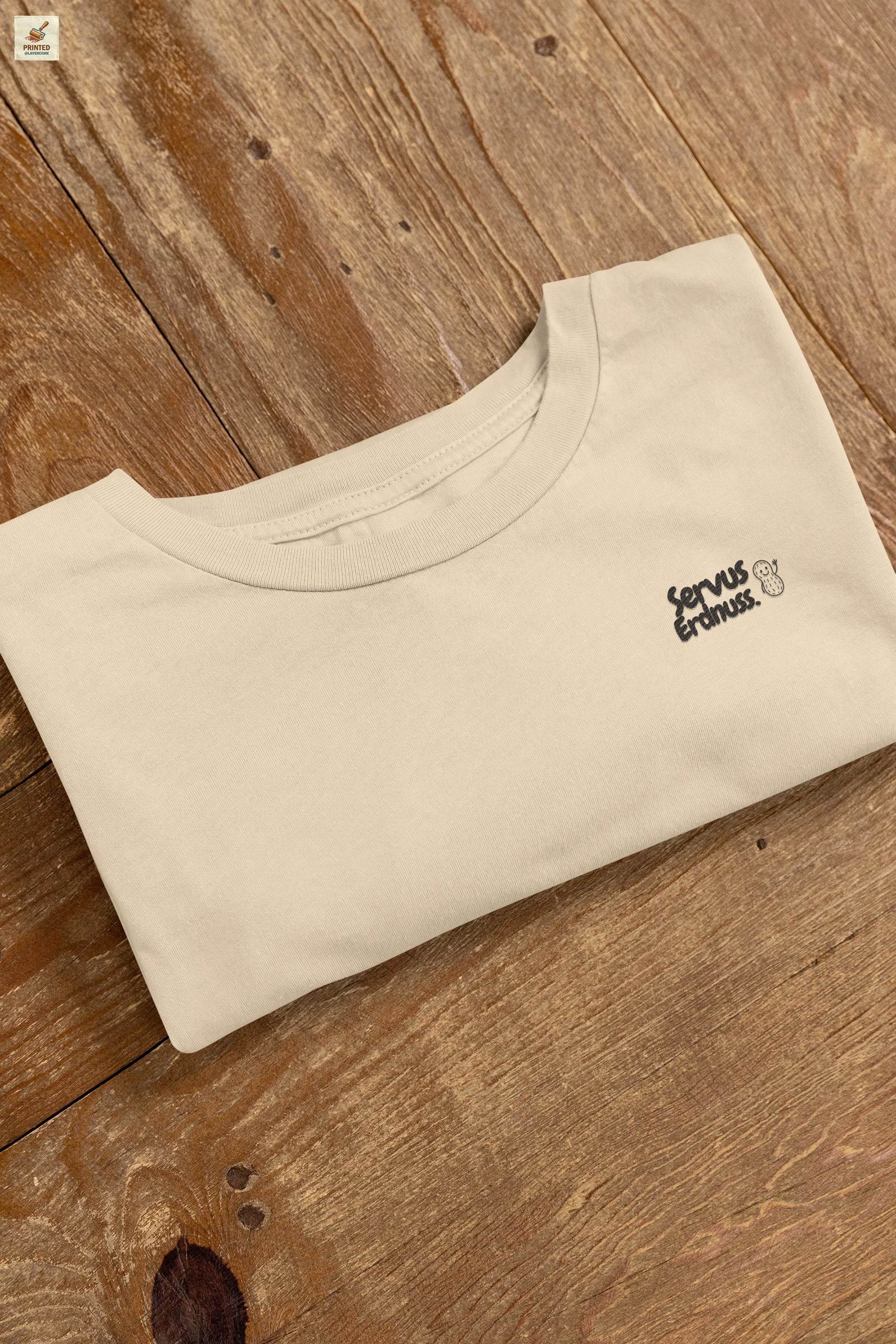Servus Erdnuss Brustmotiv mit Dialekt-Text und kleiner Erdnussfigur auf naturell farbenem Shirt