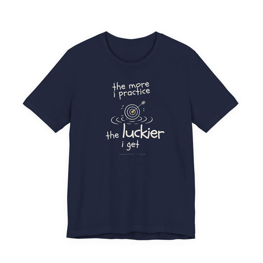 Minimalist motivational T-shirt design shown on a dark blue shirt: hand-drawn cream lettering reads “the more i practice” above a sketched bullseye with an arrow and small mustard center, ripple lines underneath, then larger text reads “the luckier i get” with tiny mono text below that says “repetition > luck.”
